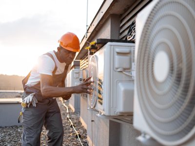 Qualified african american engineer looking at hanging air conditioner on rooftop of residential building. Competent craftsman holding modern tablet and installing device step by step outdoors.